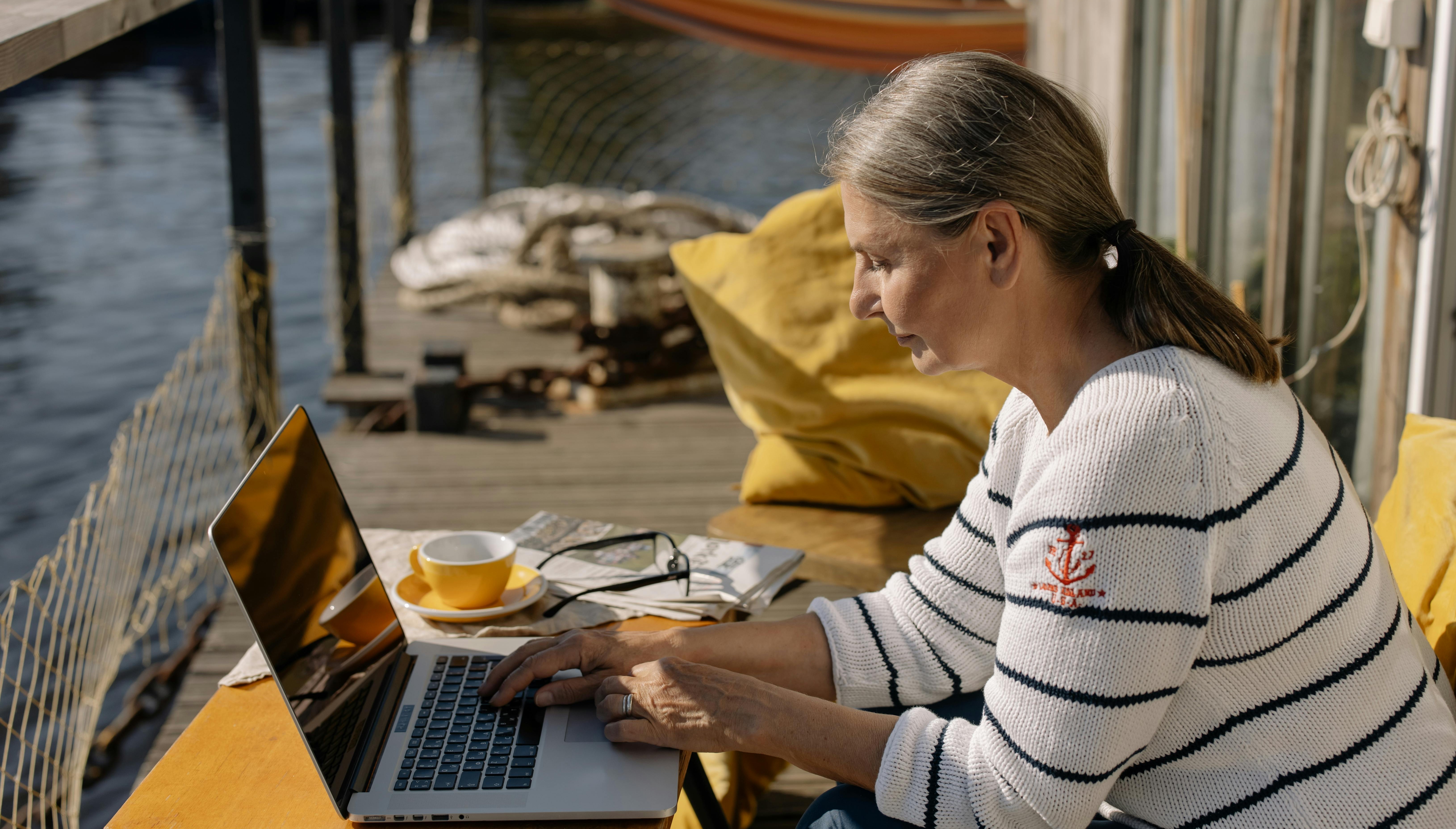 Dame sidder ved sin bærbare computer ved vandet med en kop kaffe